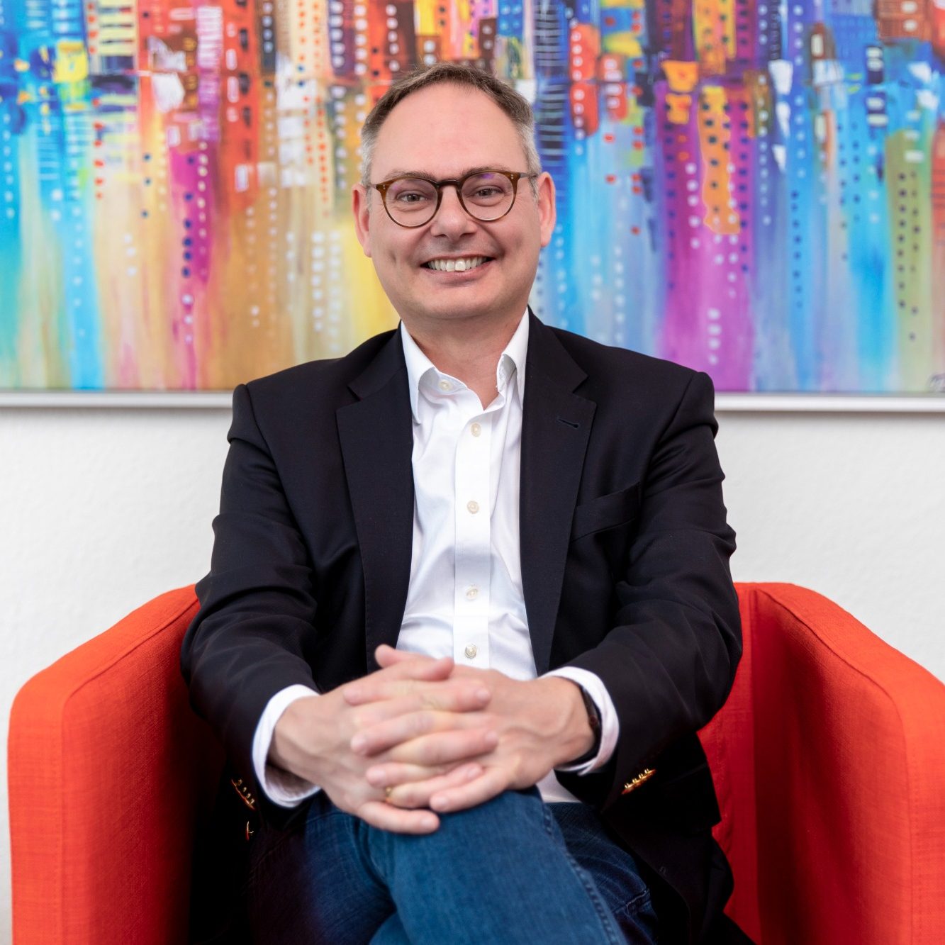 Lächelnder Mann in Brille sitzt entspannt auf einem roten Sessel vor buntem Hintergrund.